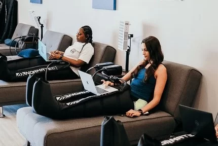 Two women enjoying Leg Compression Treatment while working on laptops at Restore